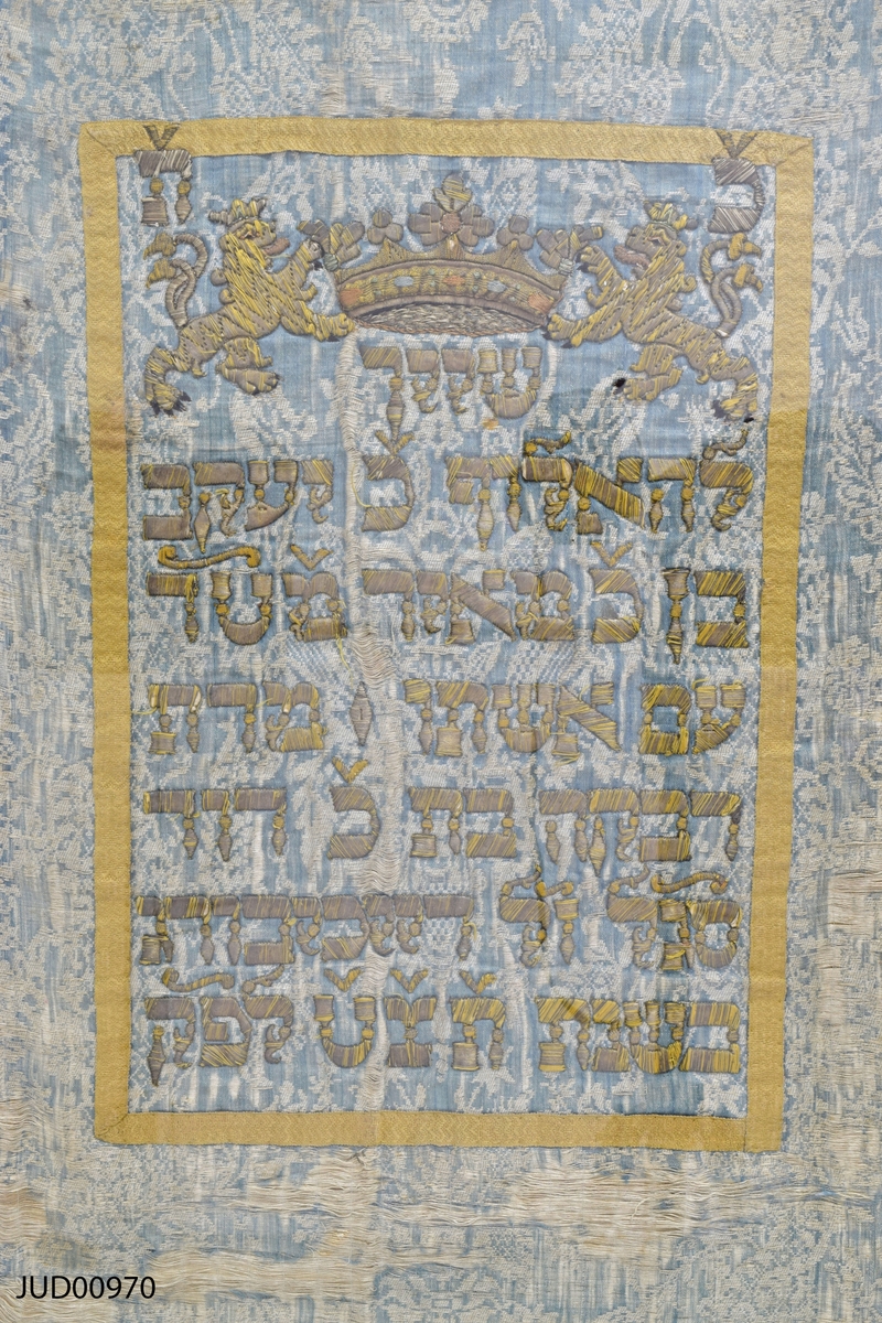 Torahförhänge i siden med röd frans nedtill och text i mitten.