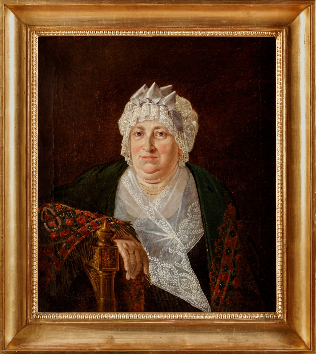 Porträtt av kvinna svept i sjal och spetsmössa på huvudet. Armen lutad mot en stol.