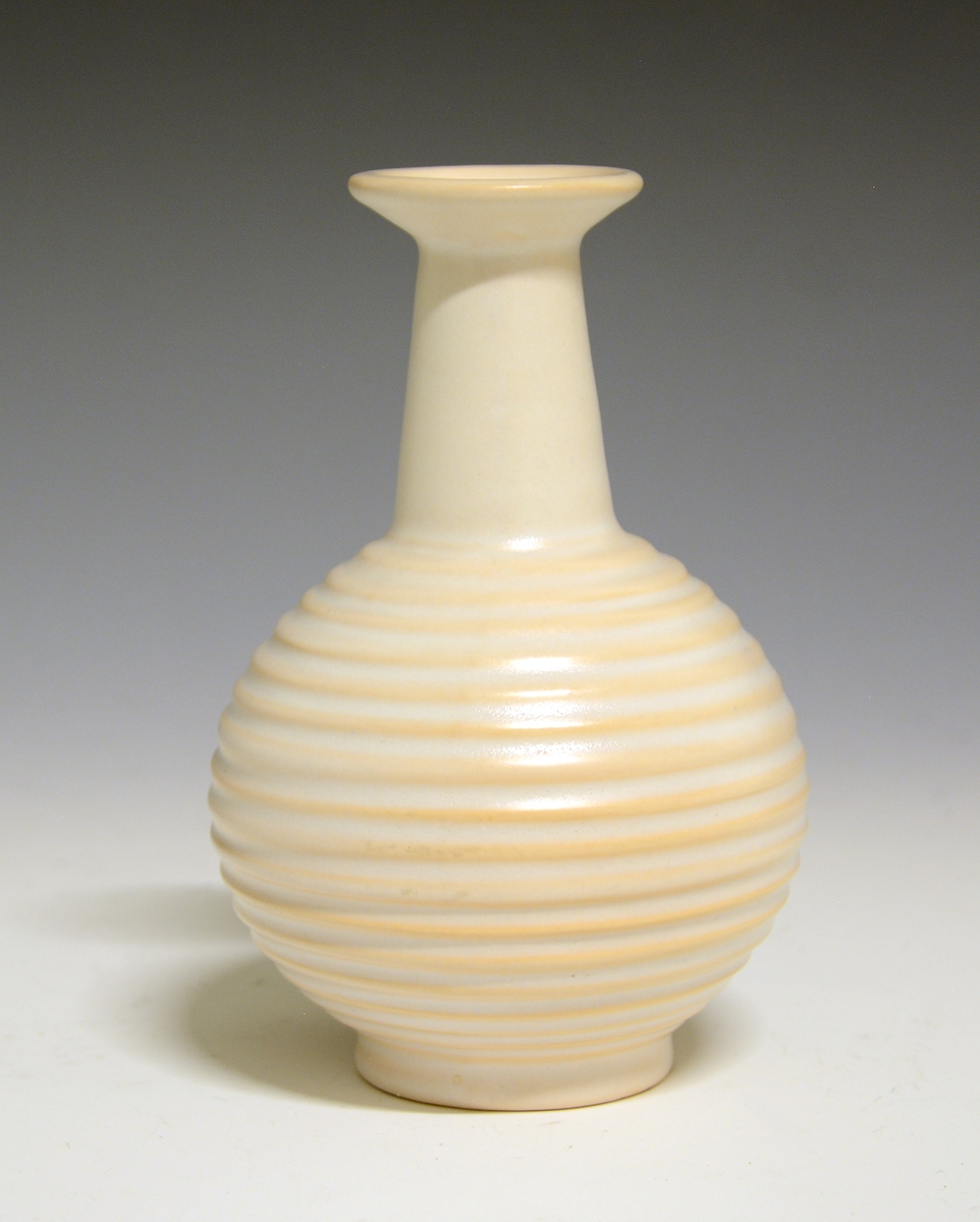 Prot: Liten vase av "keramikk". Kuleform med lav fotrand og traktformet smal hals, som er glatt mens kulen har horisontale riller. "Lyserødbeige" glasur.