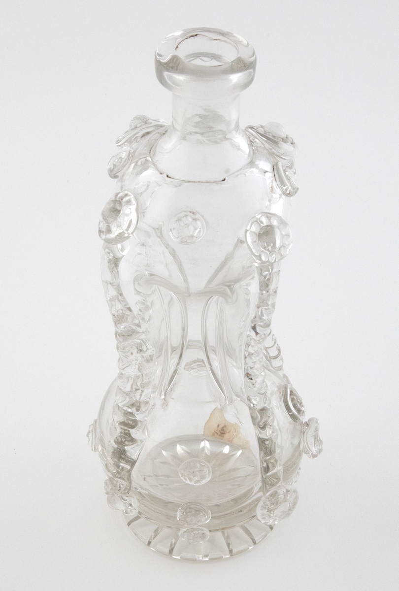 Timeglassformet klukkflaske i klart glass, hvor over- og underdelen er forbundet med et tynt rør. Korpus er dekorert med pålagte mønstre av strimler og "bringebær". Lav hals med glatt kantring, samt sirkulær fot med stjernesliping.