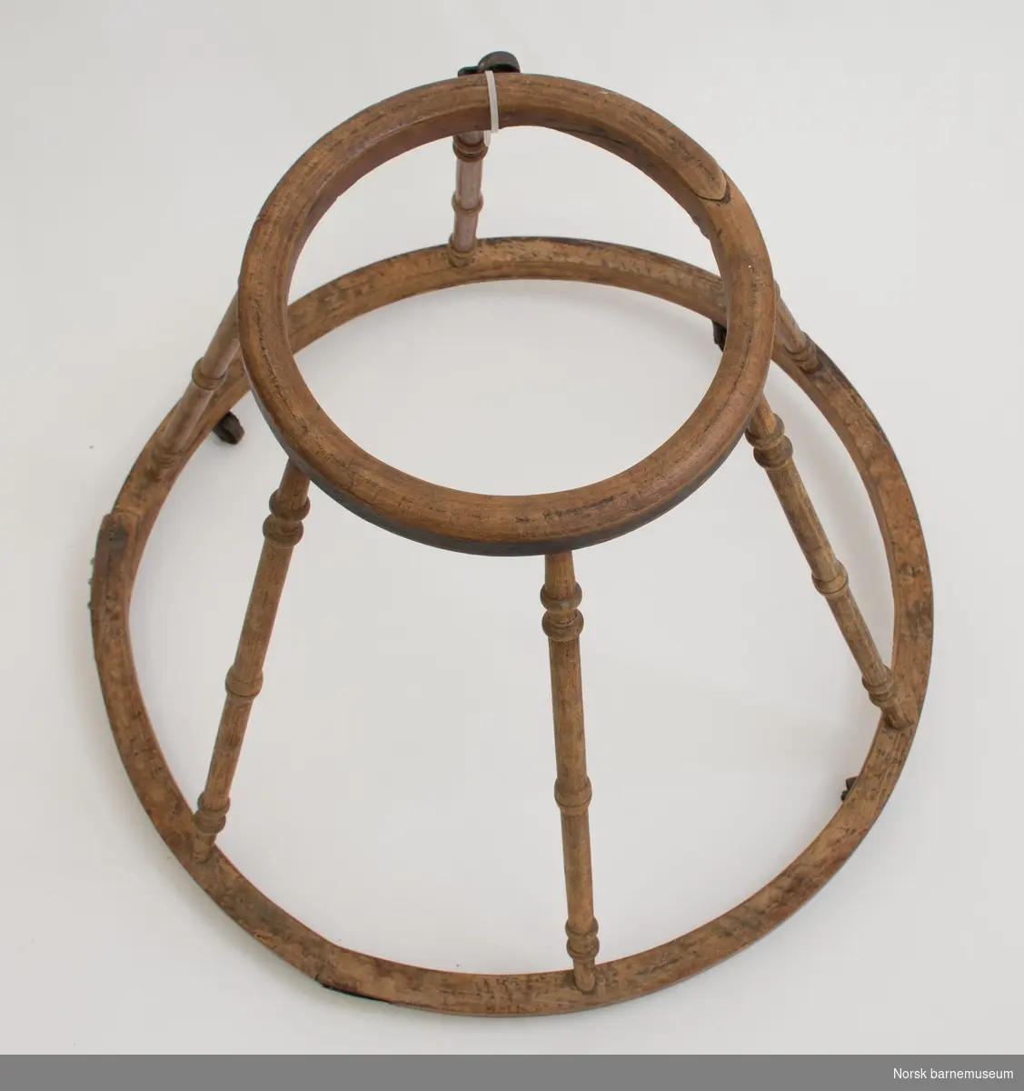 To formbøyde ringer og seks dreide spiler i bøk som står på fire hjul av jern.
Øverste ring har en diameter på 27 cm og nederste ring har diameter på 59 cm. Ett hjul er falt av. Dette følger med. Reperasjon i nederste ring