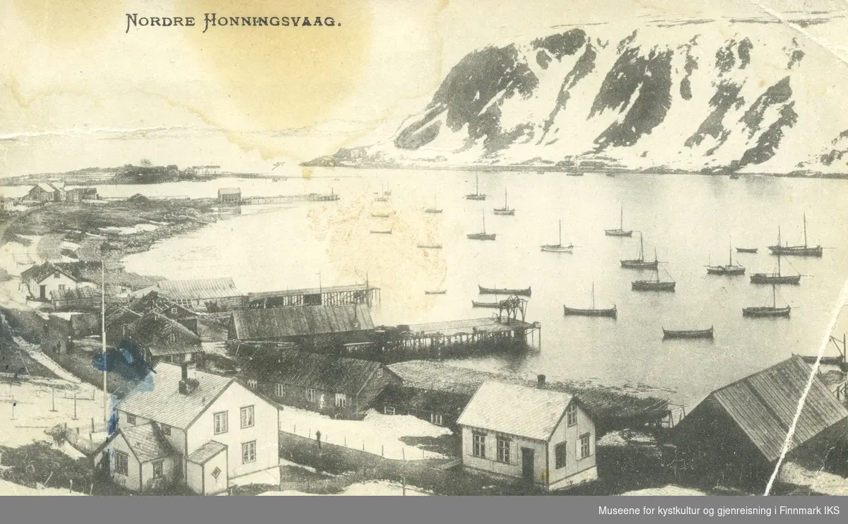 Postkort. Nordvågen. Deler av bebyggelsen og fiskebruk med kaier. Holmen med bebyggelsen i bakgrunnen. Mange Nordlandsbåter i sjøen. 1907/1908.