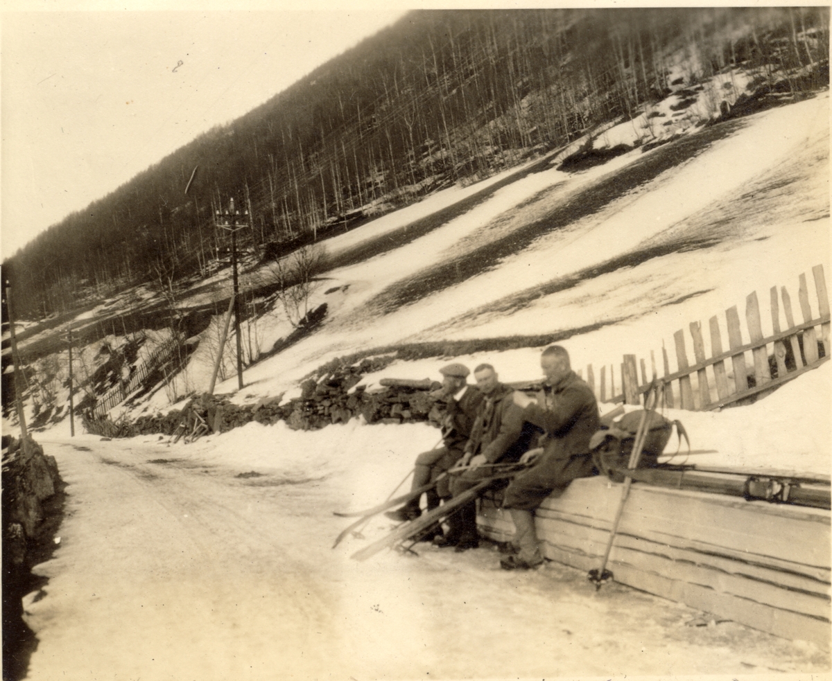 Tre deltakere i skitur fra Fokstua til Otta hviler ved en landevei på ukjent sted. Fotografert påsken 1923.