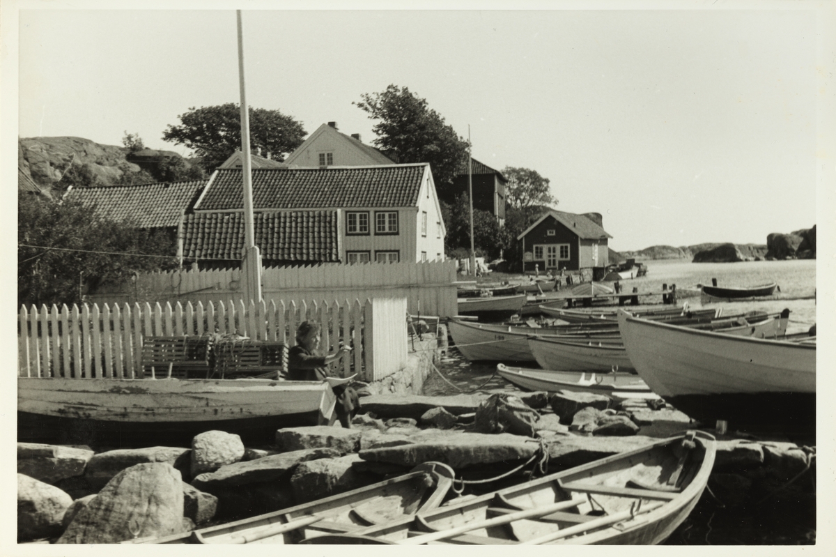 Hus og båter, antagelig i Brekkestø på Sørlandet. Foran det hvitmalte stakittet skimtes en malende Lilly Egeberg med skissebok. Fotografert sommeren 1953.