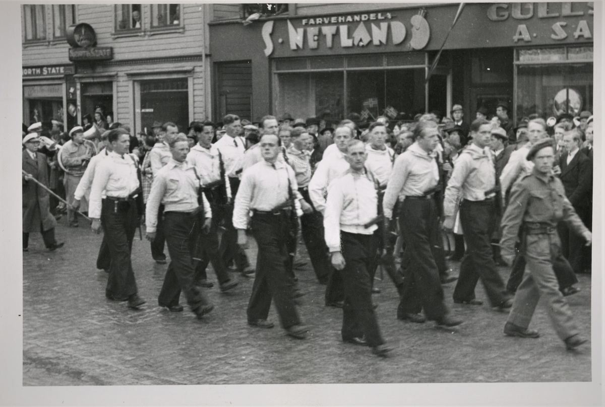 Hjemmefrontstyrker deltok i borgertoget om ettermiddagen 17. mai 1945. Her marsjerer Milorg-styrker fra Sunnhordaland i Haraldsgata forbi farvehandel S. Netaland. Musikkorps deltar i toget bak Hjemmefrontstyrkene.