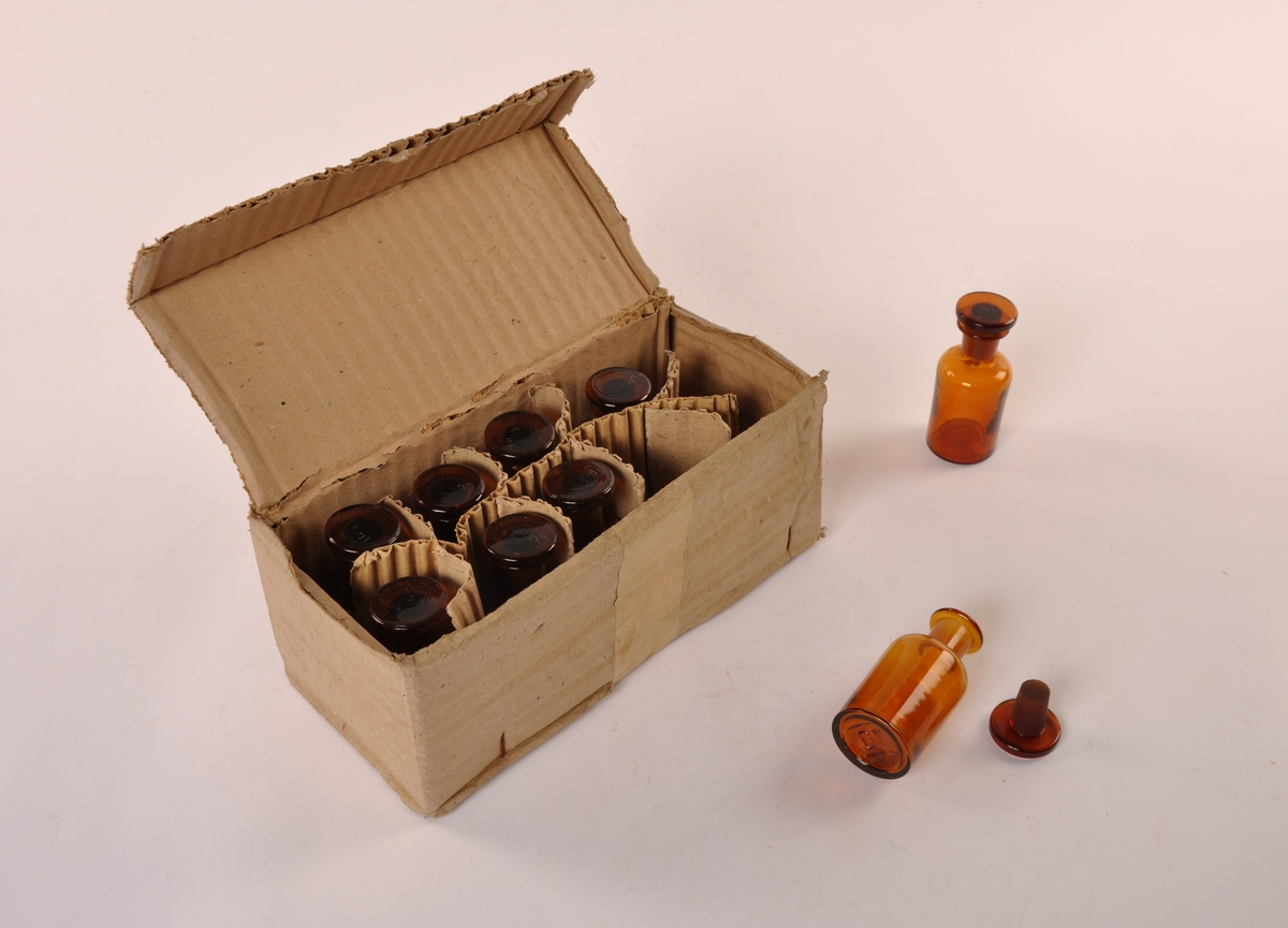 9 Stk sylindriske apotekerflasker av brunt glass med løs glasspropp. I original embalasje av papp, med etikett.