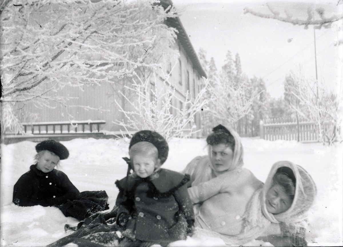 De fire døtrene til bestyrer Rydgren ved A/S Kistefos Træsliberi ligger eller sitter i snøen i hagen tilhørende bestyrervillaen. I bakgrunnen er den store arbeiderboligen på Kistefos. De yngste jentene har kåper og luehatter, mens de toeldste har hatt og sjal over hatten.