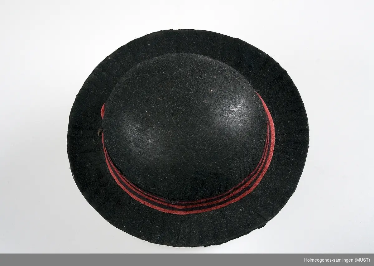 Dukkehatt av papp, sort  med grå innside. Hatten har "bowler"form. Hatten har hattebånd laget av rød og sortstripet kreppapir. Utsiden ser "filtet" ut.