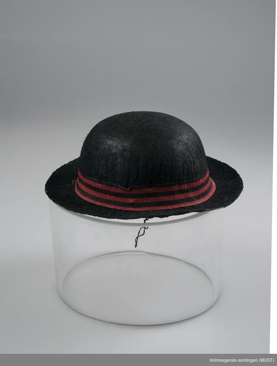 Dukkehatt av papp, sort  med grå innside. Hatten har "bowler"form. Hatten har hattebånd laget av rød og sortstripet kreppapir. Utsiden ser "filtet" ut.
