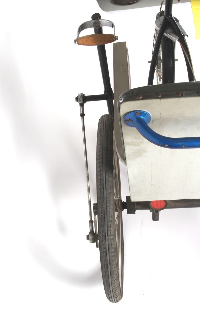 Sortlakkert invalidvogn på tre hjul med benstøttebrett og stol. Trommelbremsenav på forhjulet med bremsegrep på styrestangen. Sykkelen er hånddrevet.