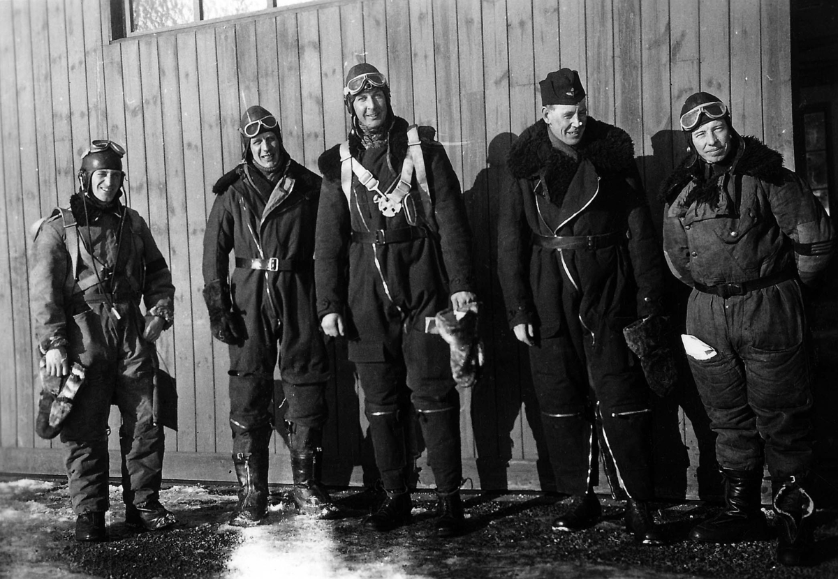 Grupporträtt av fem militära flygare i flygdräkt framför en hangarport, vintertid. 1930-tal.