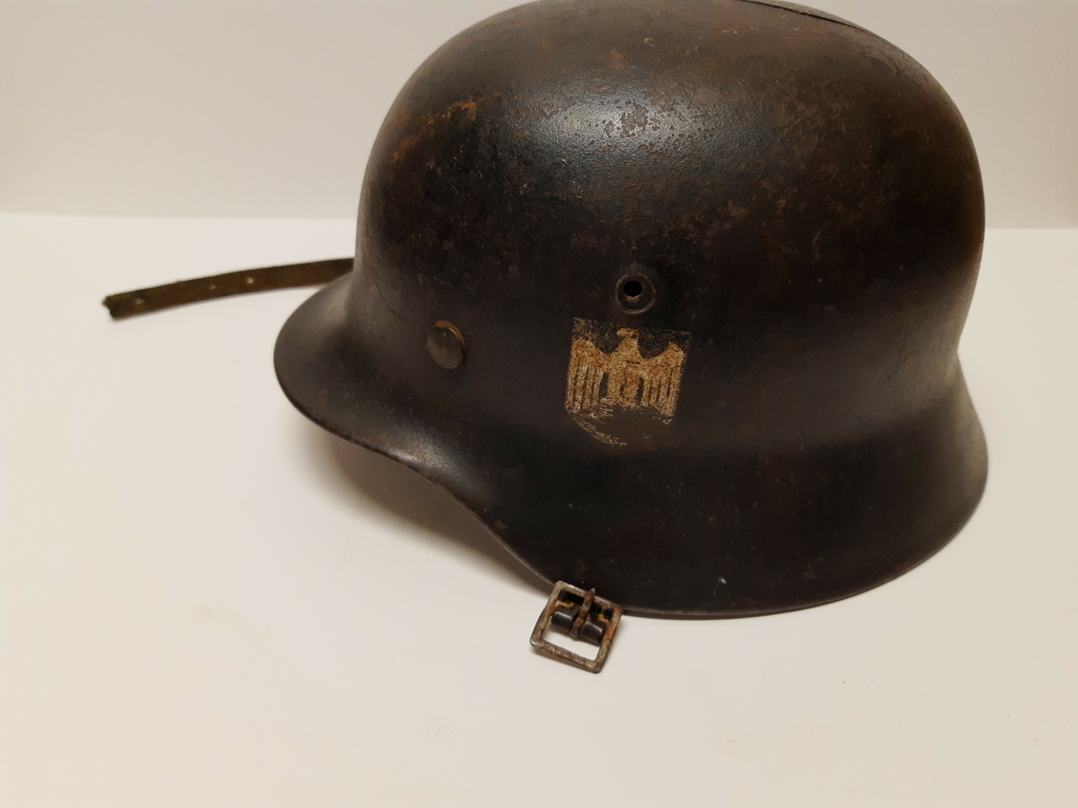 Stålhjelm av typen brukt av tyske soldater under 2. verdenskrig. Hjelmen har rem med spenne til å feste under haken. På venstre side er hjelmen dekorert med riksvåpenet til det tredje riket, ørnen over et hakekors. Merket er veldig slitt.