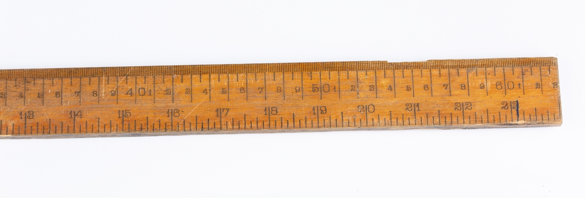 Flat, smal målestav med inndeling i cm og tommer. Lengden er lik én alen.