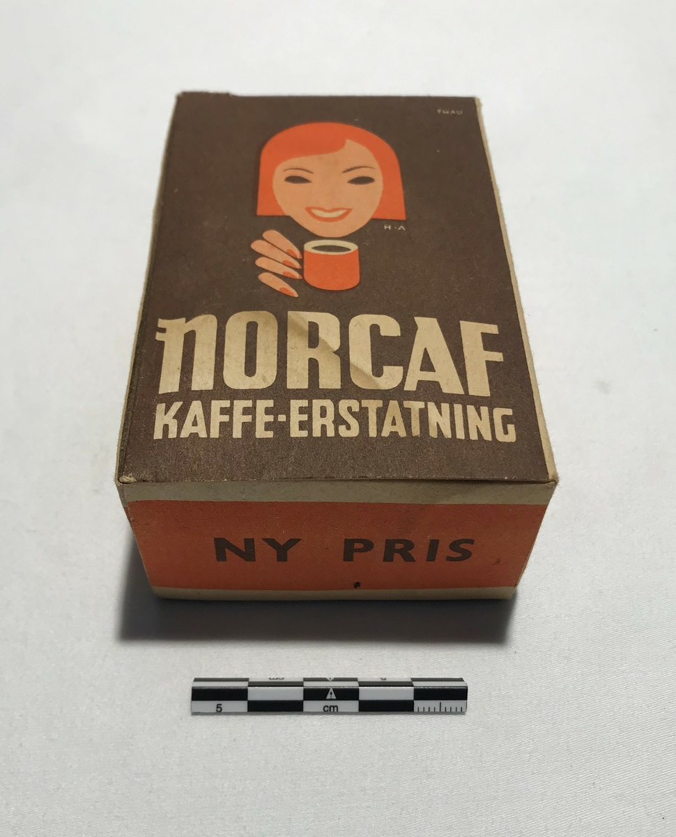 Eske av papp med kaffeerstatning. Tekst: NORCAF KAFFE ERSTATNING. På sidene står bruksanvisning på tilbereding for koking og trakting. Kaffeerstatningen ligger i esken i en papirpose.