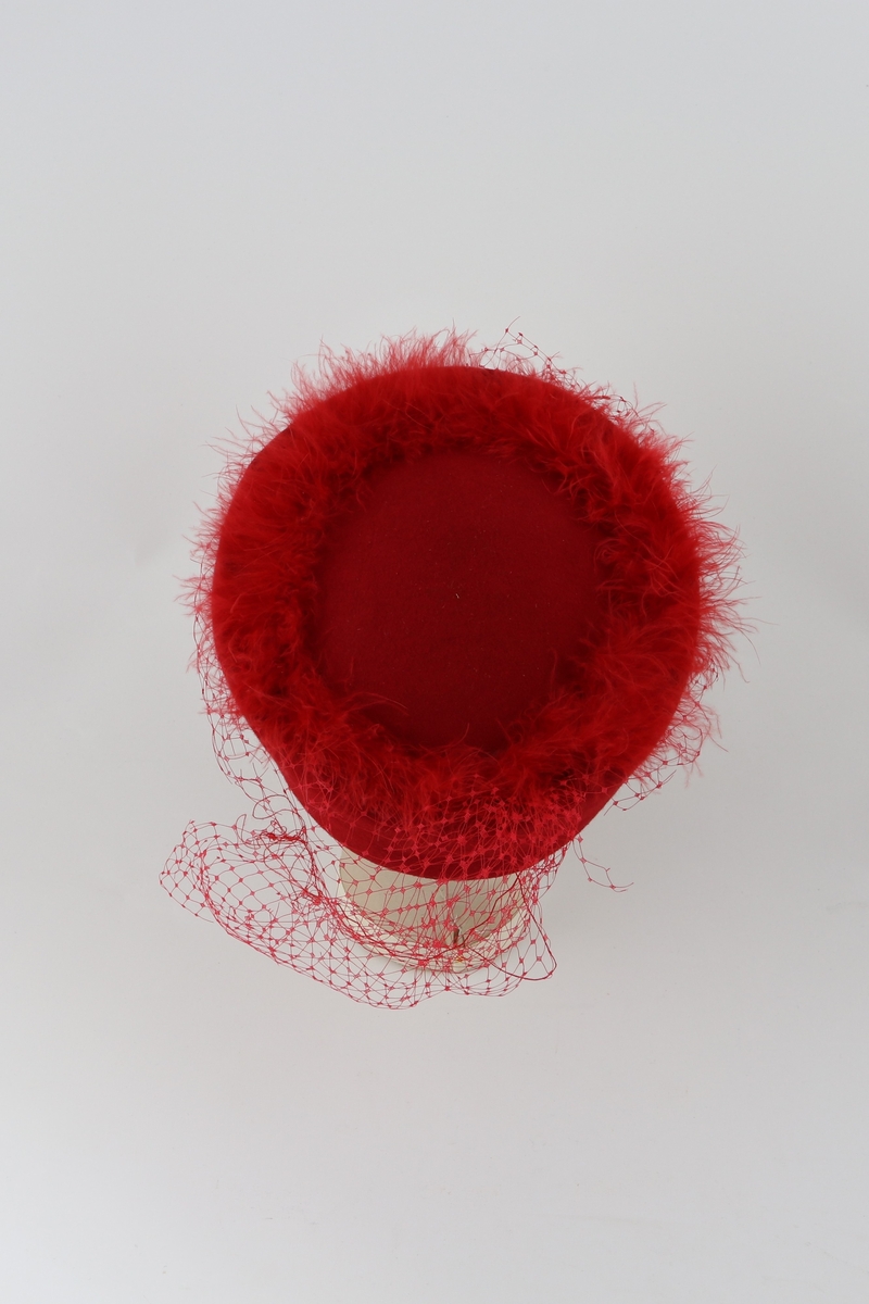 Rød sirkulær hatt i stiv filt. Bord av syntetiske røde fjær rundt hattens topp. Rød netting festet under fjærene henger ned på sidene og ved bakparten.