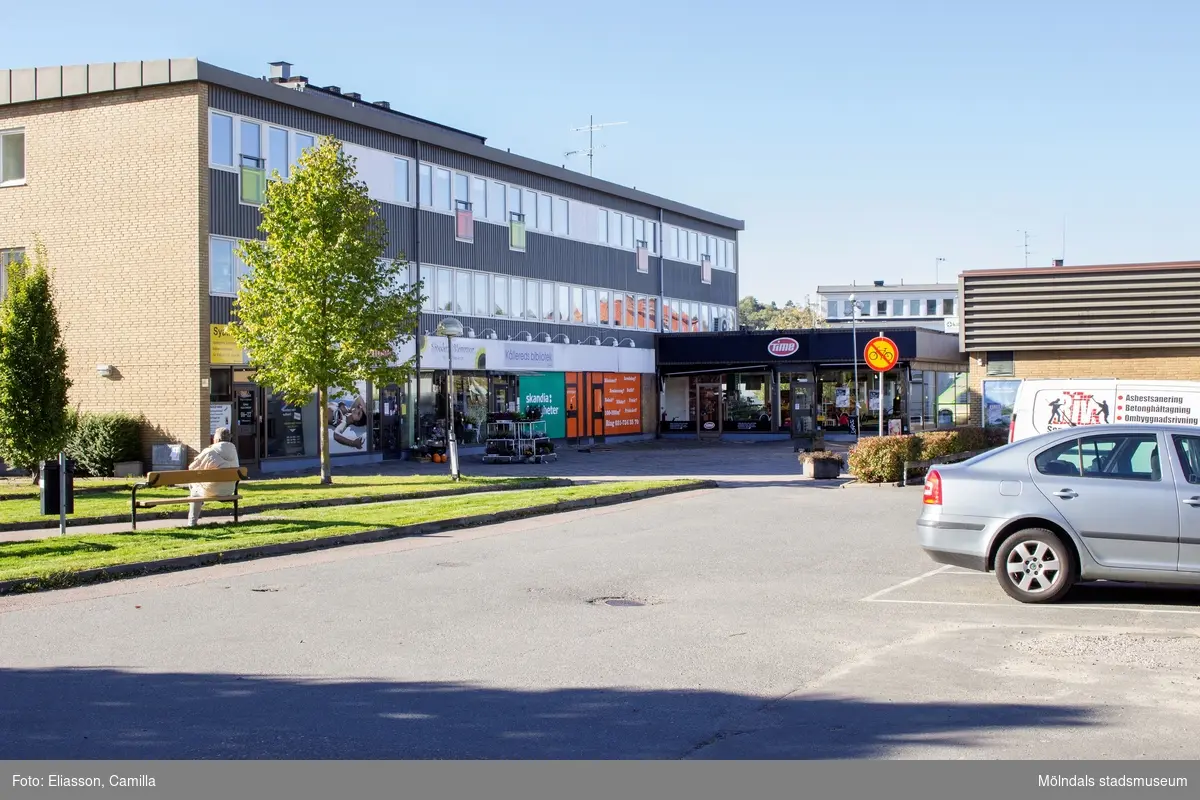 Vy västerut från Våmmedalsvägen, över en parkeringsplats, mot det inre torget i Kållereds centrum den 6 oktober 2016. Affärsbyggnaden till vänster är Hagabäcksleden 1-13. Byggnaden inrymmer bland annat en syateljé, Solhuset Kållered, Sivedens blommor, Kållereds bibliotek och en Time-butik. Byggnaden till höger har adress Gamla Riksvägen 67.