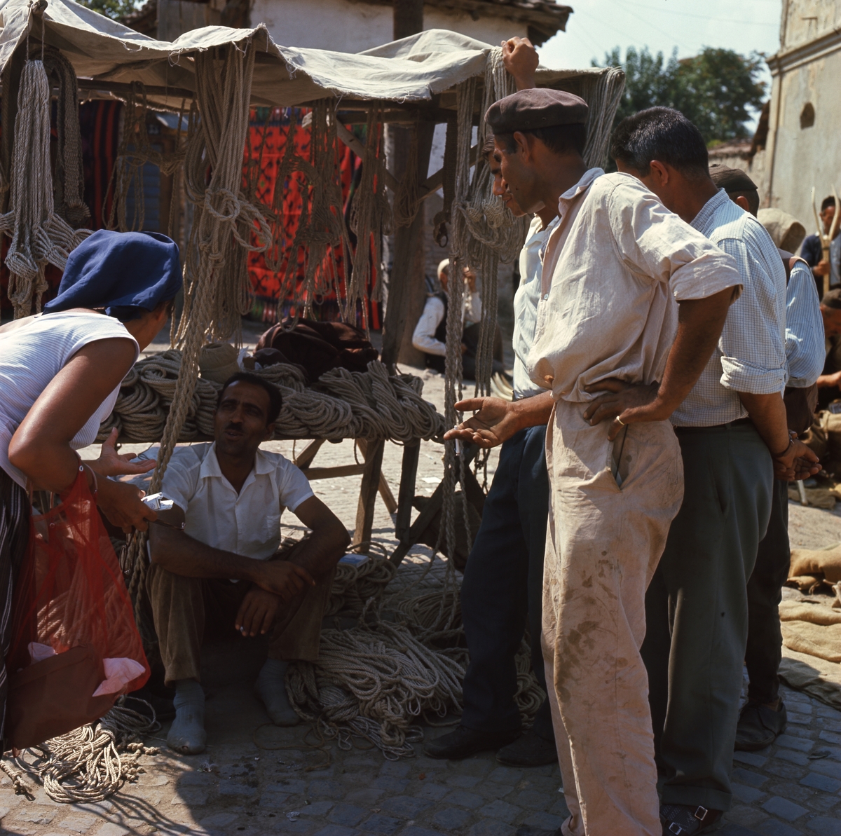 Framför ett marknadsstånd med rep sitter en man på marken. Framför honom står en kvinna och flera män.