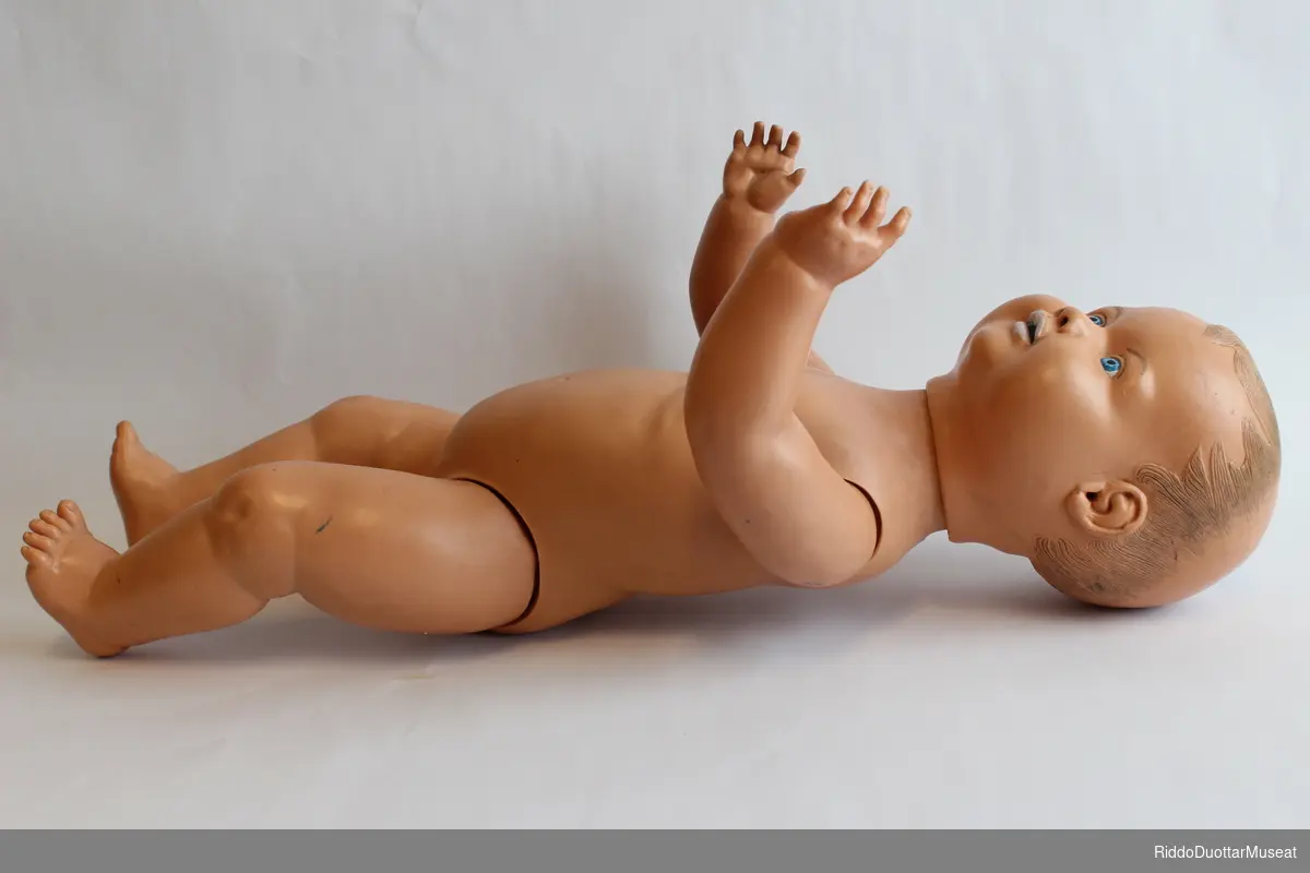 Hardplast dukke med bevegelige armer og bein. Hodet er tungt og faller bakover og til siden. Dukken skal blant annet kunne brukes til å lære hvordan man må holde babyens hode ved løft.