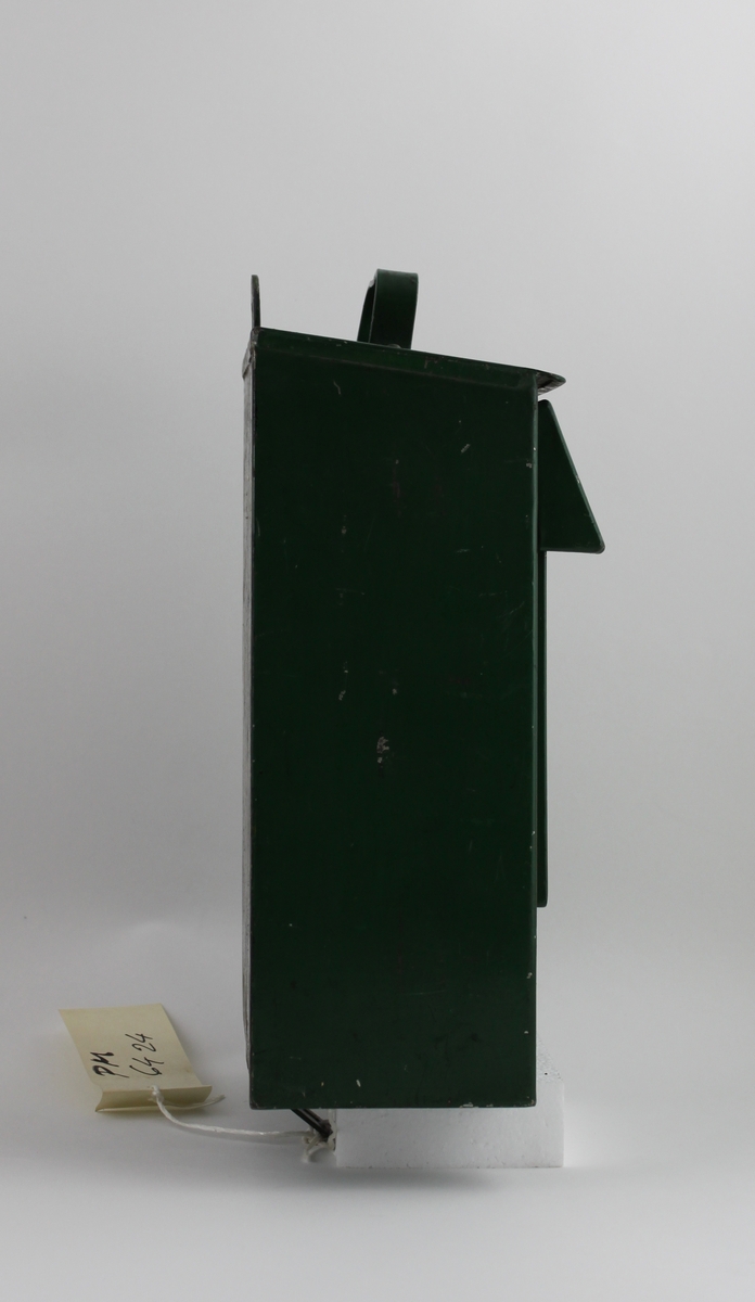 Brevlåda, med fronttömning, på klaffen över brevinkastet finns ett målat sigillförsett kuvert. På brevlådans tak finns ett handtag.