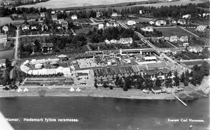 Flyfoto som viser tivolitelt til venstre og en stor plass med utstillingsboder samt Ridehuset på Hamar og omliggende villabebyggelse. Mjøsa i forkant.