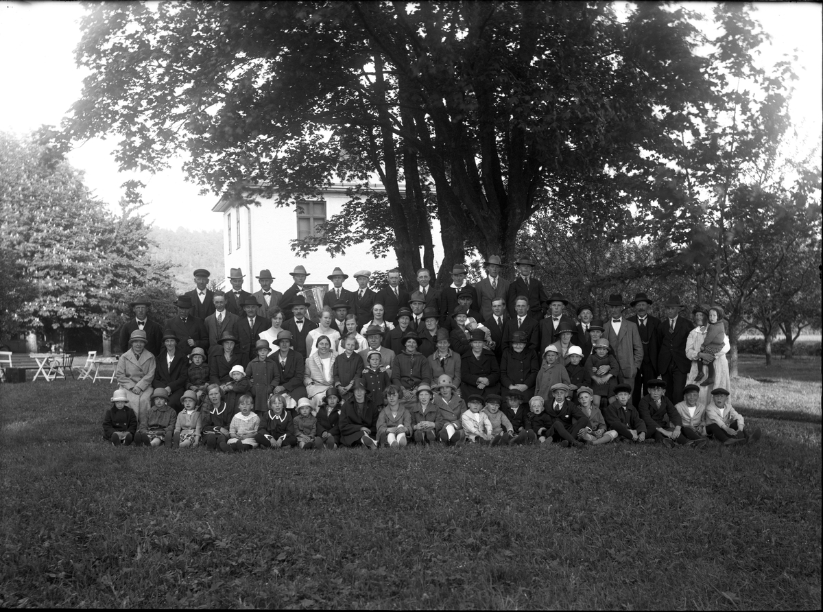Grupp med kvinnor, män och barn samlade på en gräsplan under ett stort lövträd.