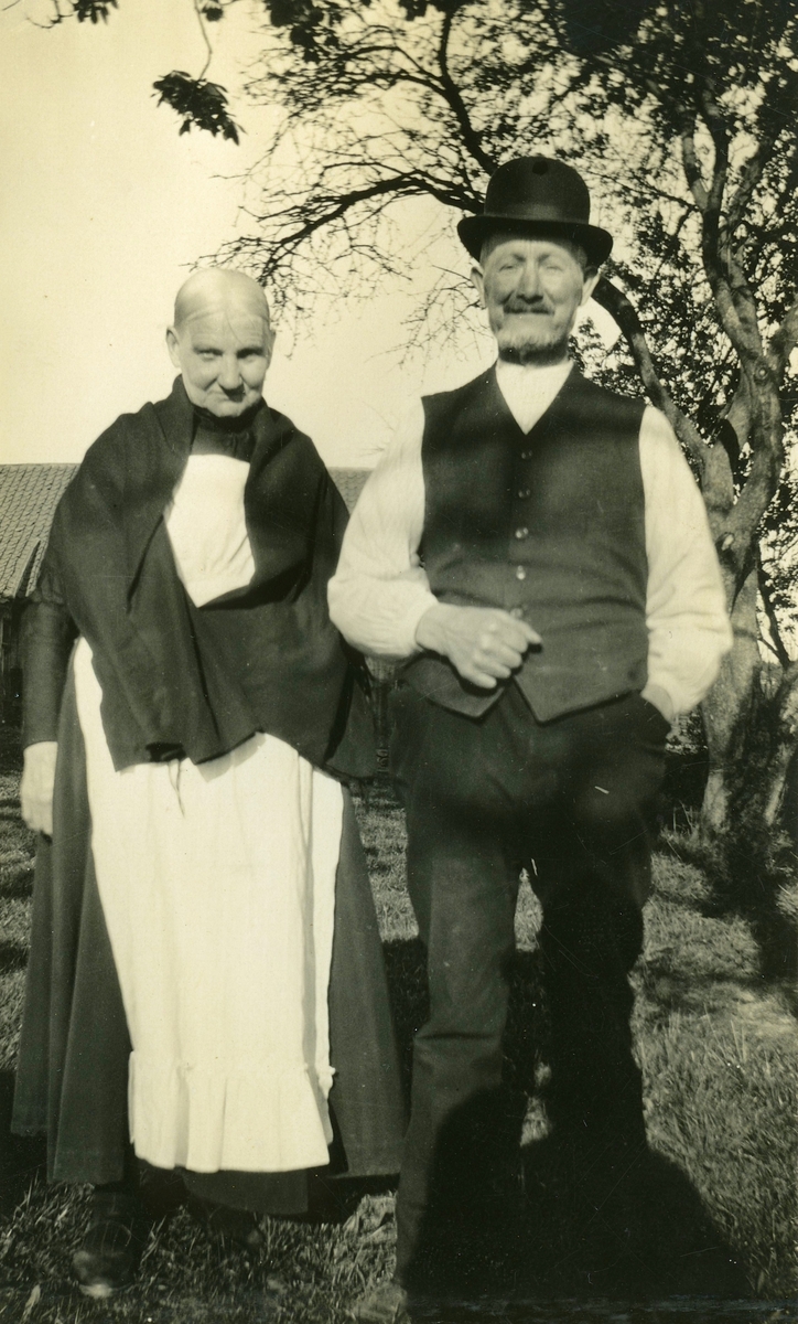Emma (född Andersdotter 1854 i Landvetter, död 1931 i Livered) och maken Karl Andersson (född 1858 i Livered "Majas", död 1931 i Livered "Majas") står utomhus snett framför ett träd, Livered "Majas" cirka 1920.