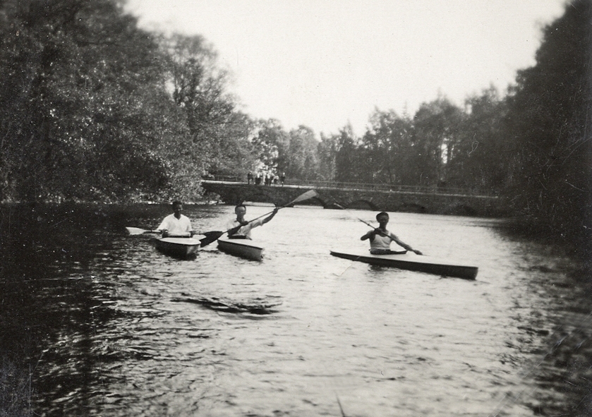 Tre kanotister paddlar kajak på en å. I bakgrunden skymtar en stenbro med några åskådare.