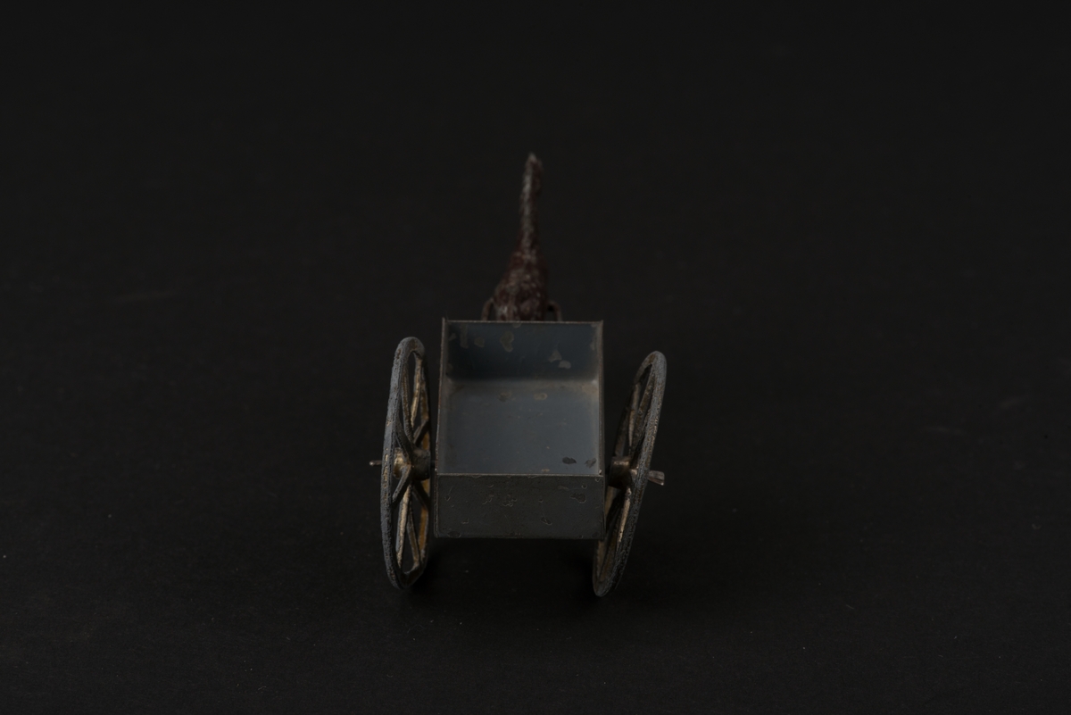 Leksak i form av häst och enaxlad vagn. 
Hästen är gjuten i tenn och brunmålad. Vagnens flak är tillverkat av järnplåt. Flaket och skaklarna är gråmålade. Hjulen var ursprungligen förgyllda.