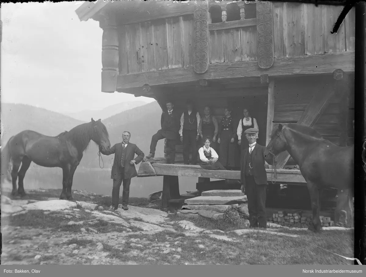 To menn holder hver sin hest foran et loft. Seks mennesker står på loftstrappa.