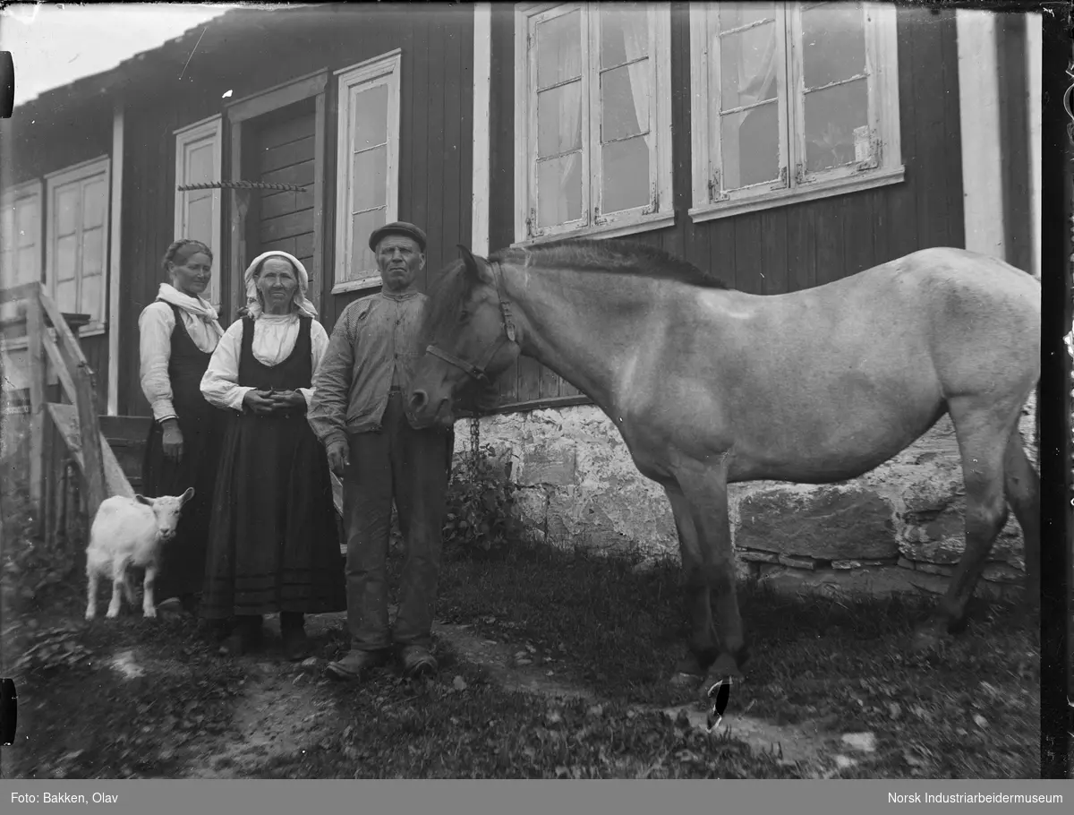 En mann og to kvinner med hest og geit foran forpakterboligen på Prestegården. Den ene kvinnen har ei rive i hånda. Menneskene er forpaktere på prestegården.