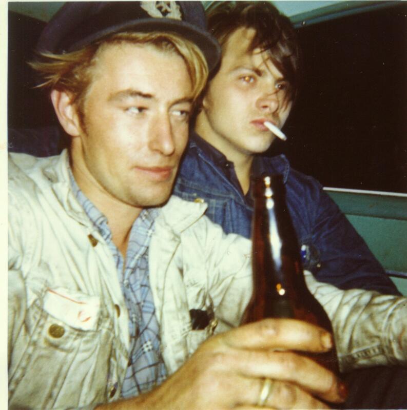 To unge menn i bil, en med brun glassflaske og en med sigarett i munnen.