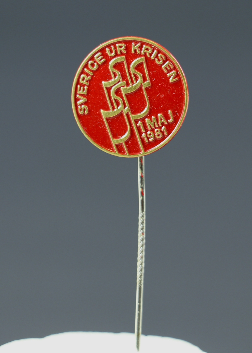 Ett runt nålmärke i guld och rött med präglad text. Längs med kanten står mot röd bakgrund "Sverige ur krisen" I mitten tre röda fanor och strax under dem står "1 maj 1981".