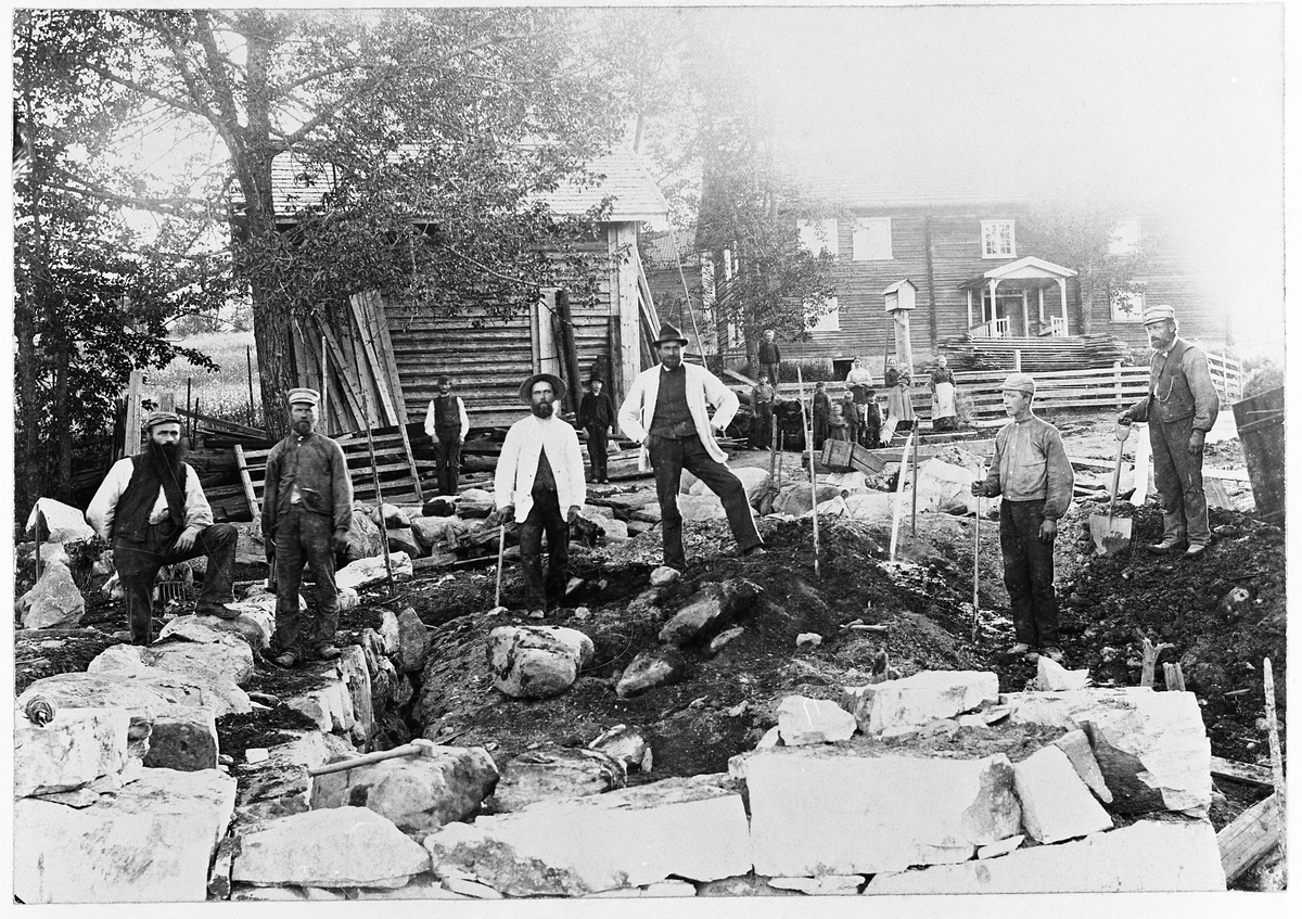 Bygging av låve i Amlien søndre, Vestre Toten, ca. år 1900.
Personene: Rasmus J. Amlie, Hans Dyrudløkken (murer), Svein Amlund, Johan J. Amlie, ukjent, Torvald Bakke.