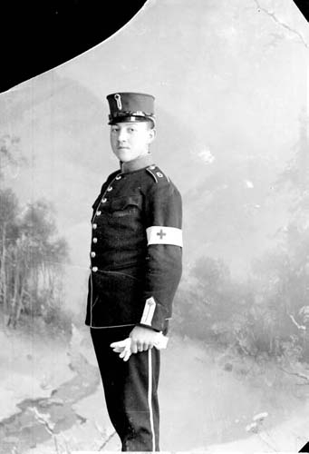 En man i militär uniform stående i 3/4-profil. En vit band med kors på vänster arm.