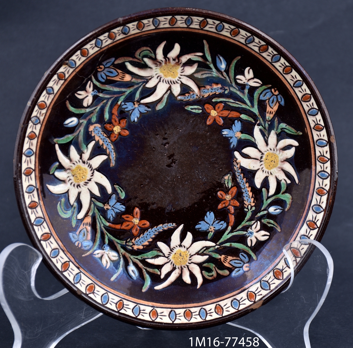 Fat (till kaffekopp) i lergods med detaljerad målad och ristad dekor föreställande blommor, bland annat edelweiss. Tillverkare är manufaktur Josef Wanzenried, verksam i kommunen Steffisburg i distriktet Thoune i Schweiz.