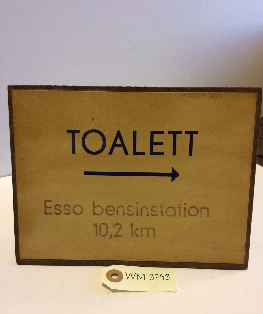 Kvadratisk skylt med text i svart på gul botten: "Toalett", en pil åt höger, samt texten "Esso bensinstation 10,2 km".