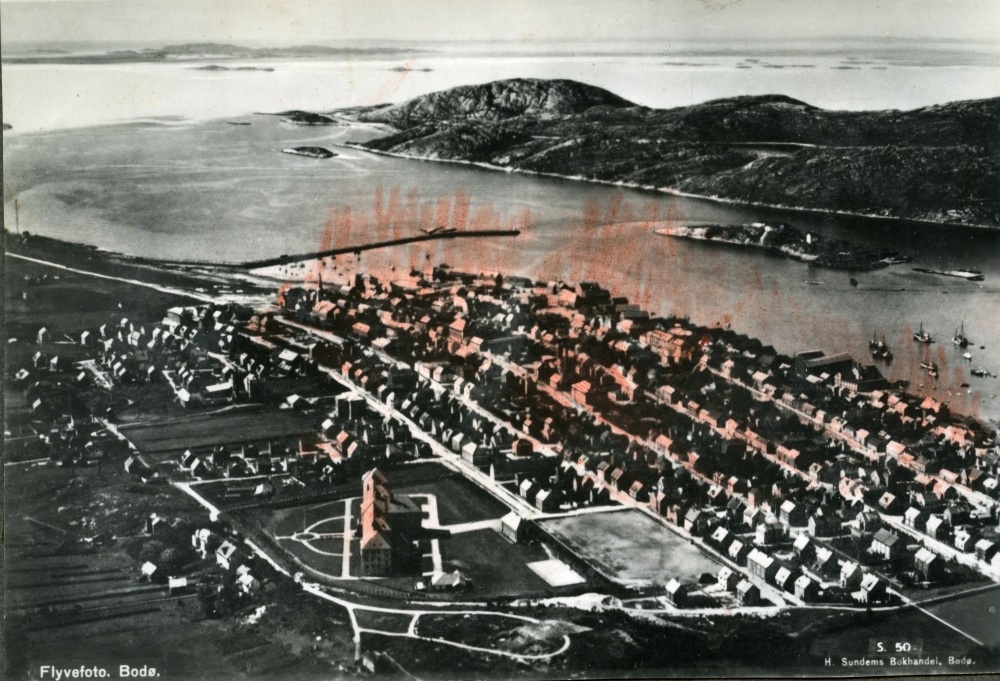 Postkort med motiv fra Bodø. Flyfoto over byens sentrum med sykehuset i forkant og moloen og Lille Hjartøya i bakgrunnen.