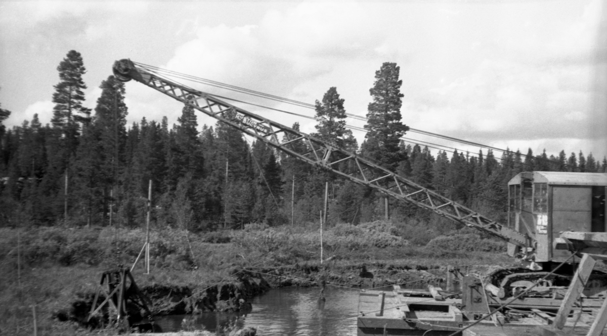 Mudringsarbeid ved Osmund, der elva Nordre Osa renner inn i Osensjøen i Åmot kommune i Hedmark. Fotografiet ble tatt høsten 1938, da entreprenørselskapet F. Selmer hadde ferdigstilt en ny dam ved Søndre Osas utløp av innsjøen. Da dammen sto ferdig startet arbeidet med å mudre, slik at regulanten skulle få utnyttet den konsesjonen de hadde fått, som innebar at myndighetene aksepterte at vannspeilet kunne variere mellom 429,50 og 437,50 meter over havet, altså en reguleringshøyde på hele 8 meter. I dette arbeidet satte man inn to maskiner. Den ene, den gang Nord-Europas største mudringsmaskin, som kunne ta inntil 6 tonn masse i hver grabb. Denne maskinen, som journalisten Per Grambo i avisa Østlendingen døpte «jutulen», ble brukt i Valmen, altså i området nærmest den nye dammen. Ved utløpene av elvene Slemma og Nordre Osa, samt i sundet mellom Vesle-Osen og Stor-Osen, i den søndre enden av sjøen, ble det brukt en maskin med mindre gripe- og løftekapasitet. Det er antakelig denne maskinen som vises på dette fotografiet - en gravemaskin som sto på en flåte. 

Mudringsarbeidene var foreskrevet av geologen Gunnar Holmsen i et utredningsarbeid han gjorde for Norges Vassdrags- og Elektrisistetsvesen i 1925, en langt mer omfattende regulering av Osensjøen enn den Christiania Tømmerdirektion hadde realisert med tanke på fløtingas behov midt på 1800-tallet. I tillegg til de mudringsarbeidene som var nødvendige ved elva Søndre Osas utløp fra sjøen, for å oppnå betydelig reguleringsintervall, foreskrev Holmsen også tiltak der hvor bygdevegen passerte Slemmas innløp i sjøen og ved Vesle-Osen, samt visse forbygningstiltak i Tverrena og Lektinga. Disse arbeidene ble tatt inn blant konsesjonsvilkårene, som ble vedtatt av Stortinget i 1928. Reguleringsarbeidene kom ikke skikkelig i gang før i 1937-38. Mudringsarbeidene skulle, i henhold til utbyggingskonsesjonen, fullføres innen utgangen av året 1940. Krigsutbruddet skapte imidlertid forsinkelser, så disse prosjektene ble ikke avsluttet før i 1941,