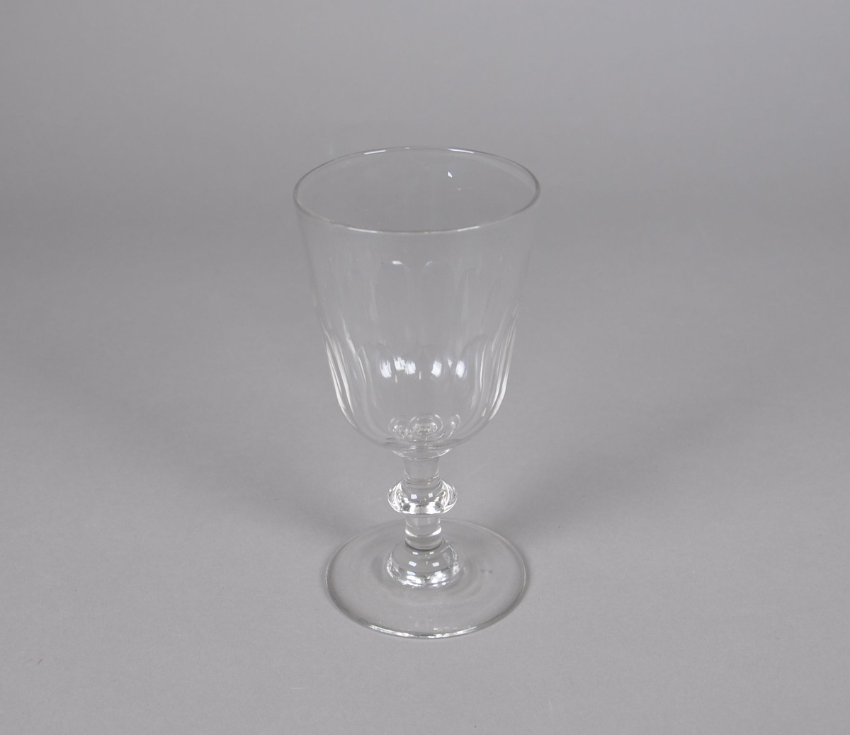 Blåst vinglass. Glasset har olivenslipt dekor på bunnen av klokken. Glasset har sylindrisk stett med dekorativ ring på midten. Glasset har rund fot.