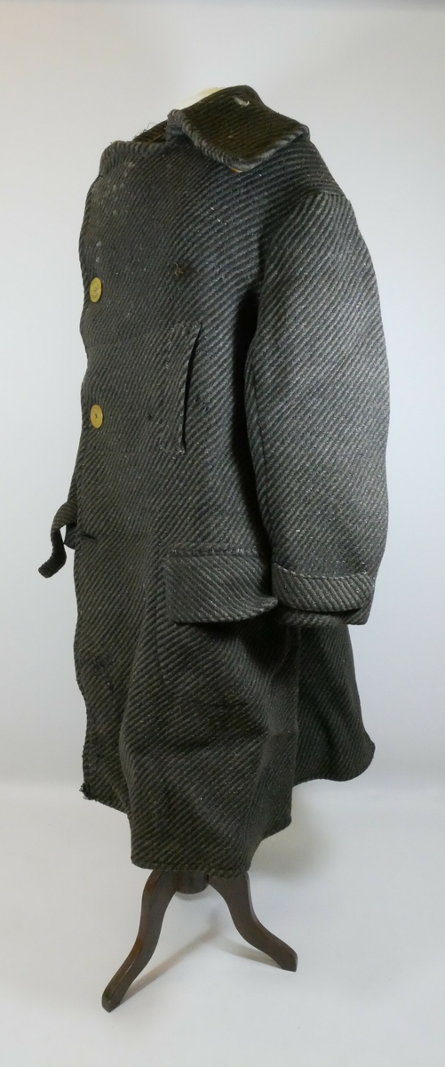 Ullkape med krage og enkeltspenn og tre gule knapper foran. En lomme på skrå og en lomme med klaff på hver side. Kåpen er stripet i grått og svart.