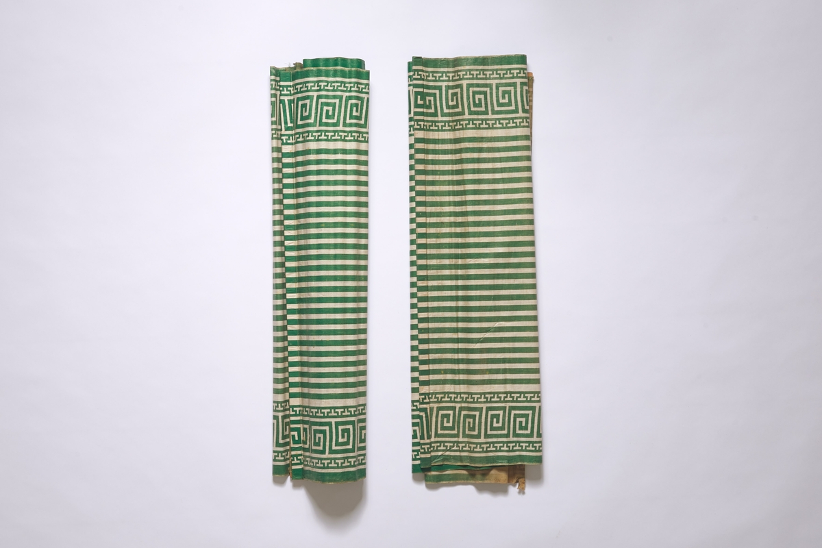 Rullgardin i lerretsvevet, hvitt stoff, med påtrykt mønster i form av grønne striper samt en meanderbord i hver side. 