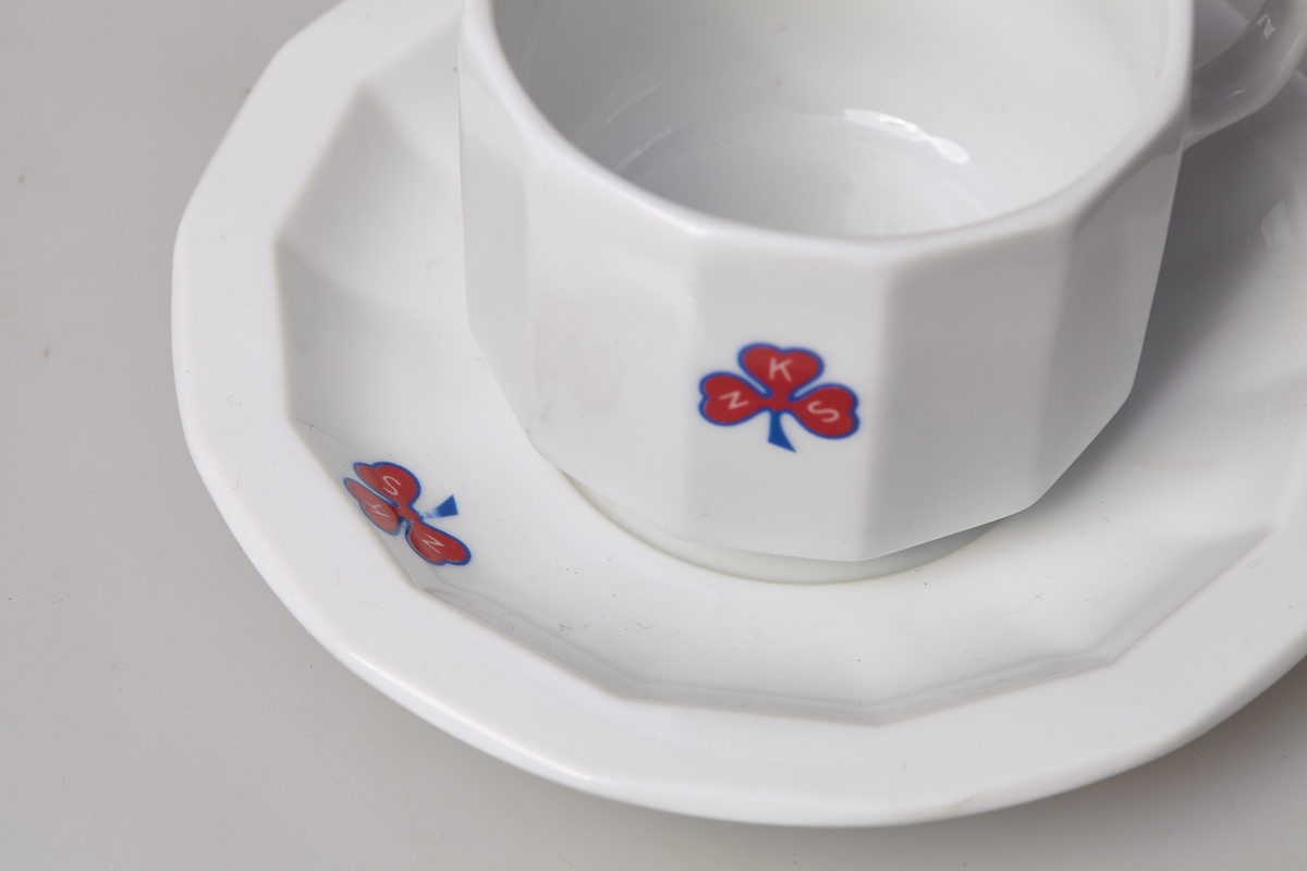Kaffekopp med skål i tykt porselen. Både kopp og skål er 12-kantet har logoen til Norske kvinners sanitetsforening. Med tilhørende asjett, TM-16060. 