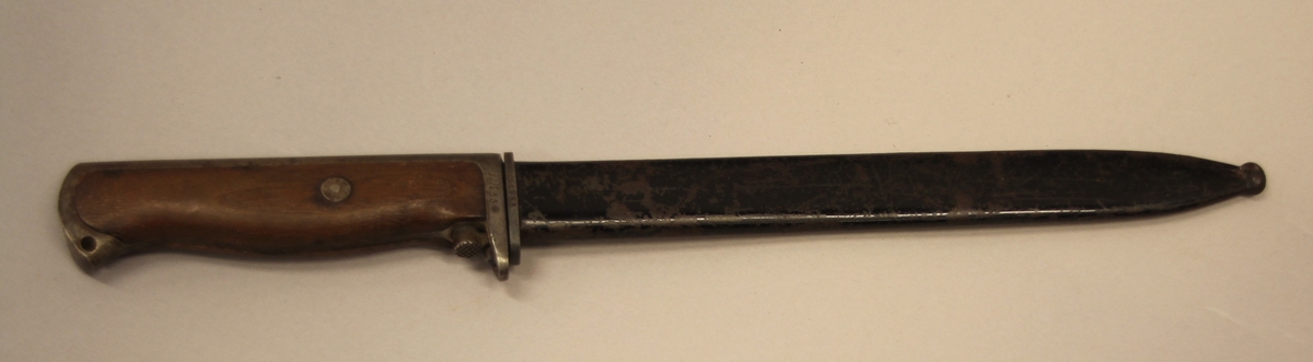 Bajonett, Krag Jørgensen mod 1912. Kongsbergmerket med 7 tal inni, viser til kong Haakon 7, og produset på Kongsberg våpenfabrikk. Bajonetten har treskaft og metallslir. Merkt 111955 både på slir og bajonett.