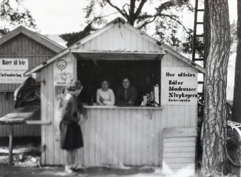 Kioskbod med to personer i luka og en kunde utenfor. Plakat til høyre for luka annonserer med utleie av båter, madrasser, strykejern og barbermaskiner. (Foto/Photo)