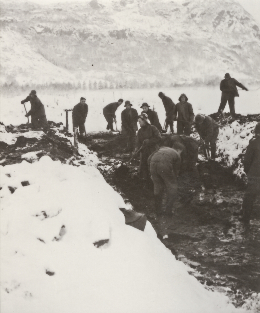 13 menn i arbeid med å grave ut en kanal. Kanalen i senter av bildet, Vinterbilde. Skogkledd åsside.