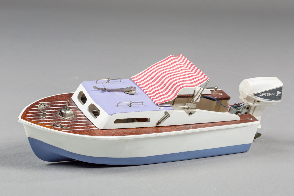Modellbåt med skrog i plast og dekk og detaljer i tre. Kalesje i tekstil. Påhengsmotor i plast som drives av batteri. Løs vindskjerm ligger oppi båten.