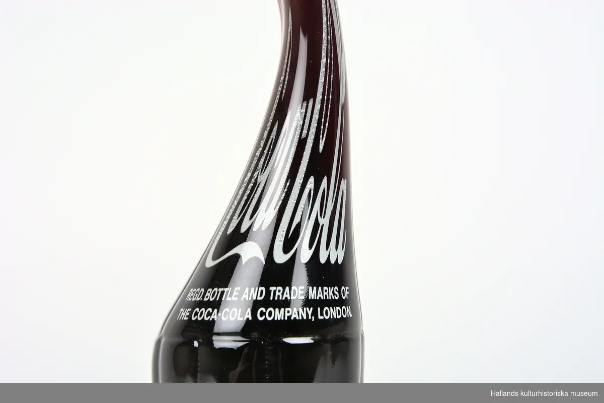 Läskedrycksflaska: Coca-Cola. Flaskan är tillverkad av klart glas och har en lång och sirlig flaskhals. 710ml. Oöppnad.