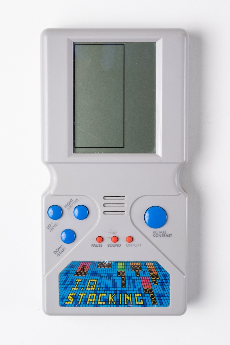 Spelkonsol, handhållen, "I.Q. Stacking" för spelet Tetris. Spelkonsolen är av grå plast med rektangulär platt form. På ovansidan finns överst en bildskärm, och därunder fyra blå knappar och tre röda knappar för styrning av spelet. Spelet drivs av 2 st batterier, AA, 1,5 V. Batterierna sitter bakom en löstagbar lucka på baksidan.