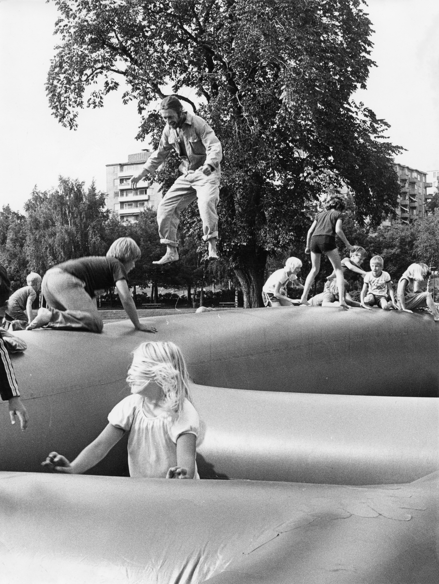 Studsmatta i Folkets Park i Linköping 1970-80-tal. Studsmatta i form av smala armar. Många barn sitter runt om på mattan, en man hoppar. I bakgrunden ser man lövträd och lägenhetshus.