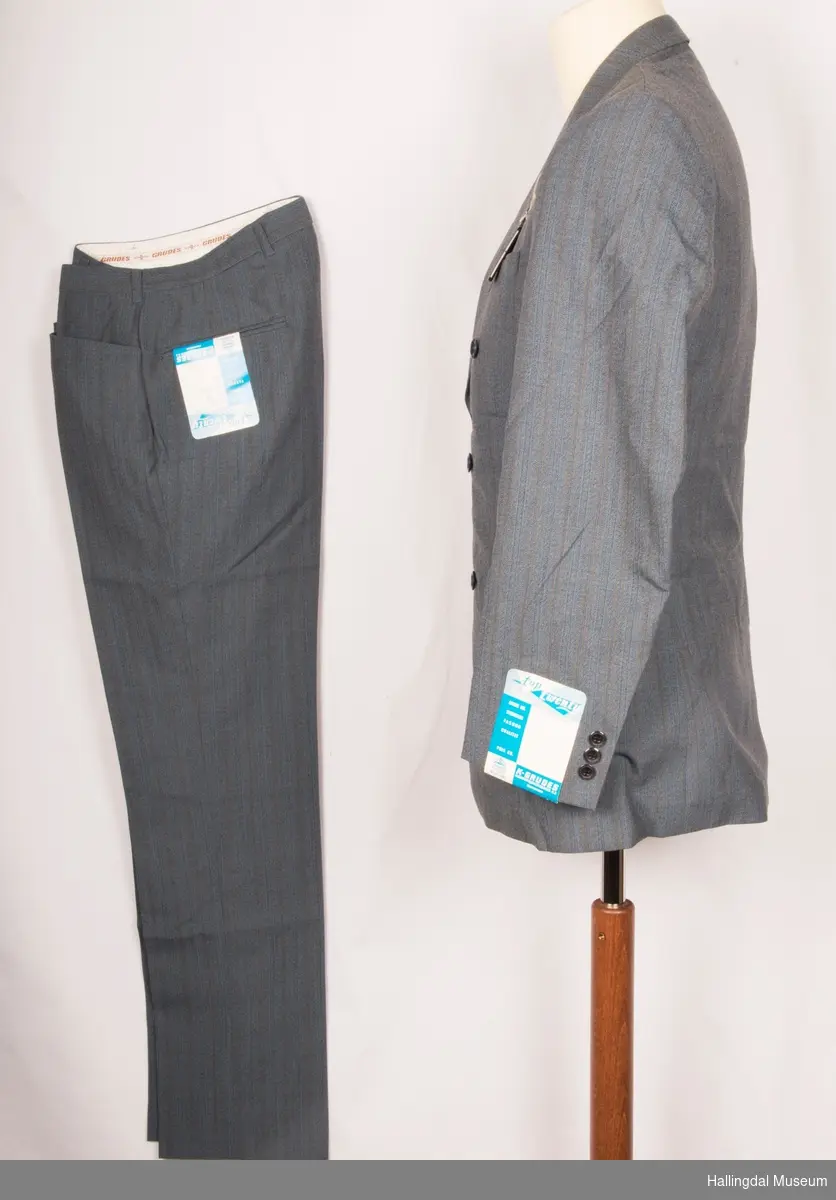 Grå herredress med jakke og bukse fra K. Grudes konfeksjonsfabrikk AS.  Dobbeltspent dressjakke med 3 knapper og knapphull på hver side.